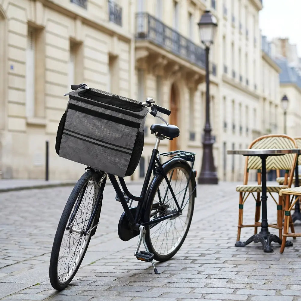 Une image de style lifestyle montrant un vélo de ville élégant équipé d'un panier de transport pour animaux gris et spacieux sur le guidon. Le vélo est garé sur une rue pavée parisienne historique, avec une terrasse de café (chaises et tables en rotin) et de superbes bâtiments haussmanniens en arrière-plan, créant une scène de transport urbain pratique et stylée pour un animal de compagnie.